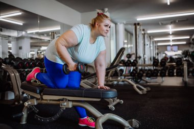 Orta yaşlı, kilolu bir kadın spor salonunda halterlerle spor yapıyor.