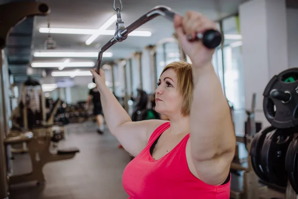 Moyenne adulte plus taille femme travaillant sur une machine lat pull-down à l'intérieur dans la salle de gym — Photo