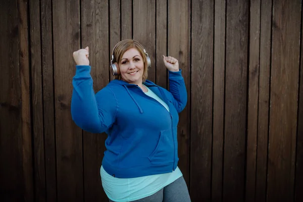 幸せな太りすぎの女性でスポーツ服とともにヘッドフォン見てカメラに対して木製の壁 — ストック写真