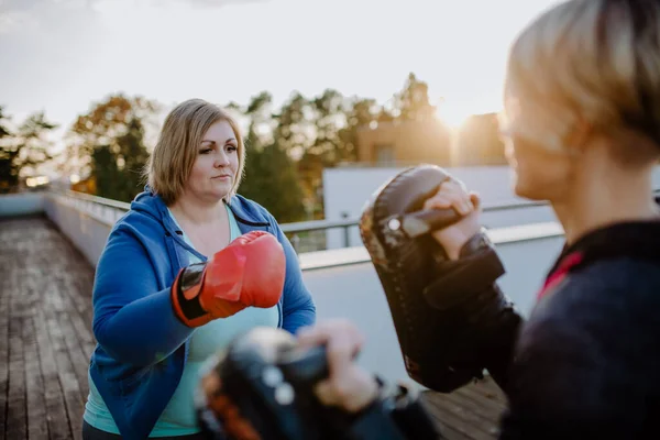 Overweight woman training boxing with personal trainer outdoors on terrace.