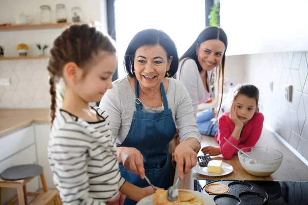 Chicas pequeñas felices con madre y abuela haciendo panqueques en casa, cocinando. —  Fotos de Stock