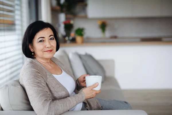 Щаслива старша жінка з кавою сидить на соамі і дивиться на камеру в приміщенні вдома . — стокове фото