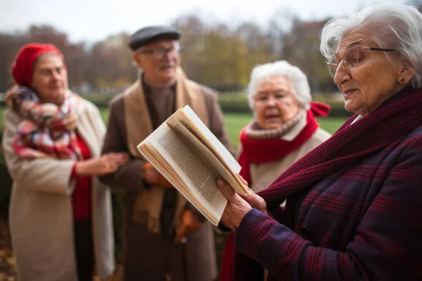 Groupe d'amis aînés heureux avec livre sur la promenade à l'extérieur dans le parc en automne, la lecture et la parole. — Photo