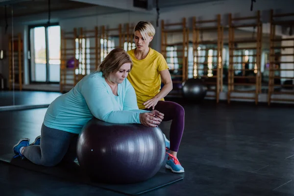 Happy overweight woman exercising with personal trainer on fintess ball in gym — стоковое фото