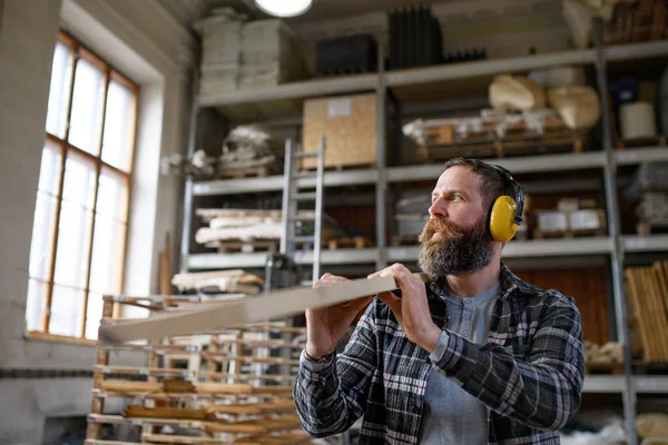 Macho carpinteiro maduro com protetores auditivos carregando placa de madeira, dentro de casa na oficina de carpintaria. — Fotografia de Stock