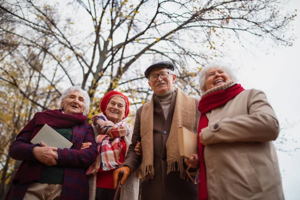 Low angle view of group of happy senior friends with books on walk outdoors in park in autumn — Foto Stock