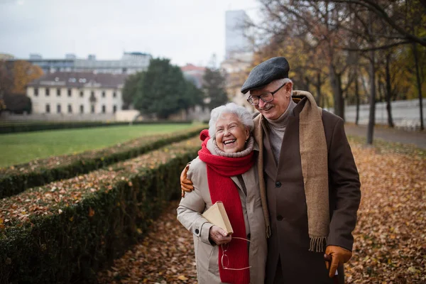 Happy senior couple on walk outdoors in town park in autumn, embracing and laughing. — Fotografia de Stock
