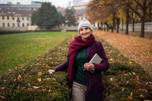 Sonbaharda kasaba parkında kitabı olan mutlu yaşlı kadın kameraya bakıyor.. — Stok fotoğraf