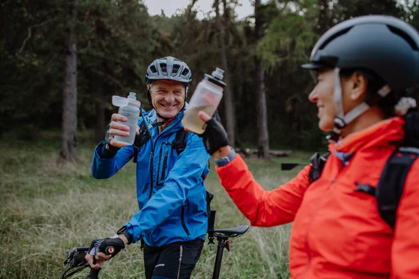 Happy senior couple bikers with wtaer bottles outdoors in forest in autumn day. — Zdjęcie stockowe