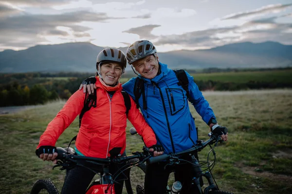 Šťastný starší pár cyklisté objímající venku v přírodě v podzimním dni, při pohledu na kameru. — Stock fotografie