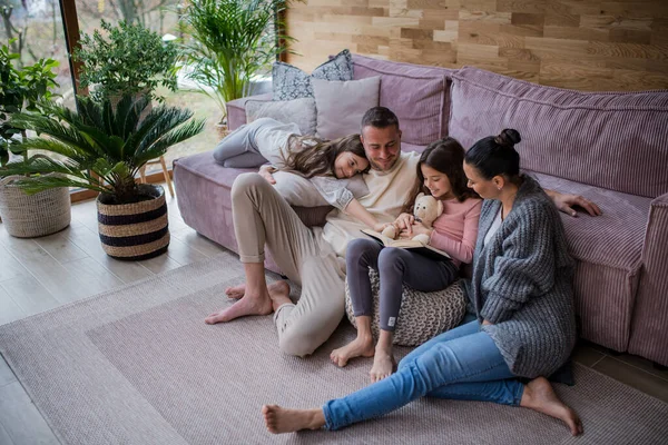 Дві щасливі сестри з матір'ю і батьком сидять на підлозі, обіймаючи і читаючи книгу вдома — стокове фото