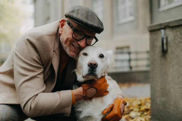 Homem sênior feliz olhando para a câmera e abraçando seu cão durante a caminhada ao ar livre na cidade. — Fotografia de Stock