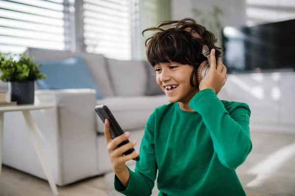 Szczęśliwy wielorasowy chłopiec ze słuchawkami i smartfonem słuchającym muzyki w domu. — Zdjęcie stockowe