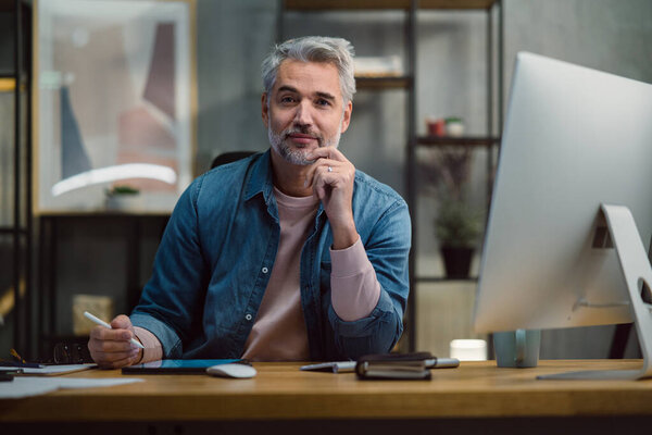 Mature man architect working on tablet at desk indoors in office, looking at camera.