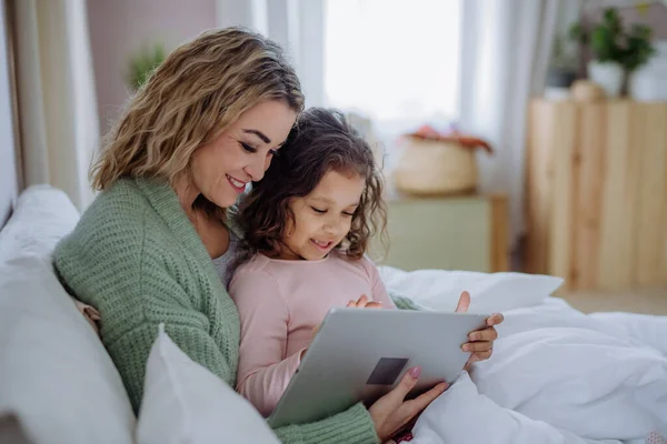 快乐的妈妈带着她的小女儿躺在床上，在家里用平板电脑. — 图库照片