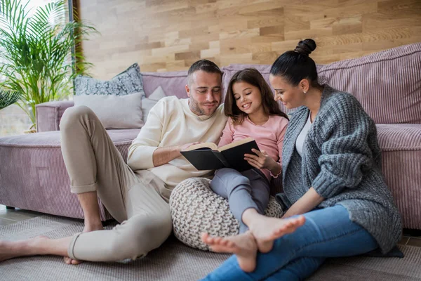 Glad liten flicka med mor och far sitter på golvet och läser bok hemma — Stockfoto