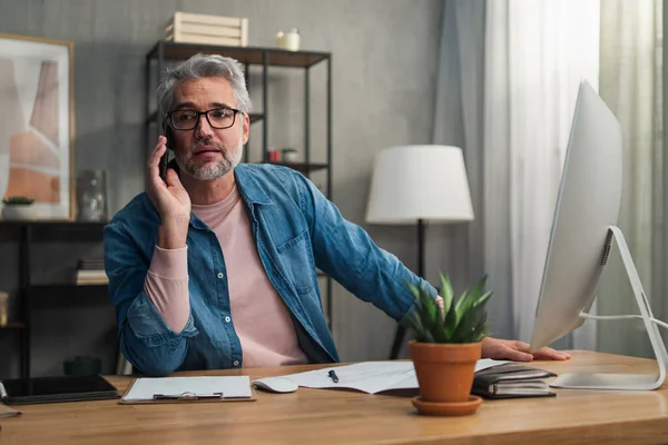 Reifer Mann arbeitet am Computer und telefoniert am Schreibtisch im Büro. — Stockfoto