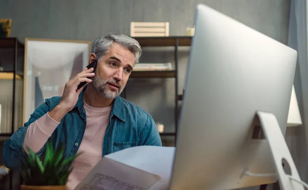 Volwassen man architect werkt op de computer en het maken van een telefoontje aan de balie binnen in het kantoor. — Stockfoto