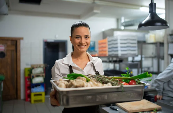 Female chef carrying tray with fresh vegetables and looking at camera in restaurant kitchen. — Stock Photo, Image