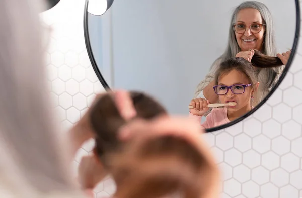 Бабушка с внучкой стоят в помещении в ванной комнате, расчесывая зубы и волосы по утрам — стоковое фото