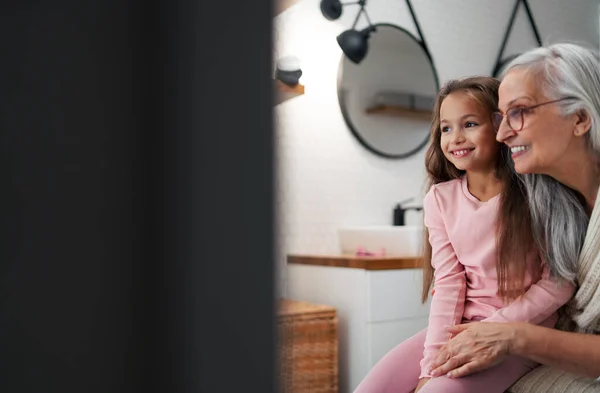 Senior grootmoeder en kleindochter staan binnen in de badkamer, knuffelen. — Stockfoto