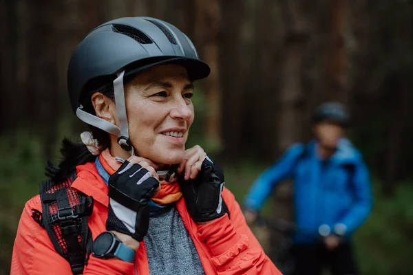 Senior žena biker uvedení na cyklistické helmy venku v lese na podzim. — Stock fotografie