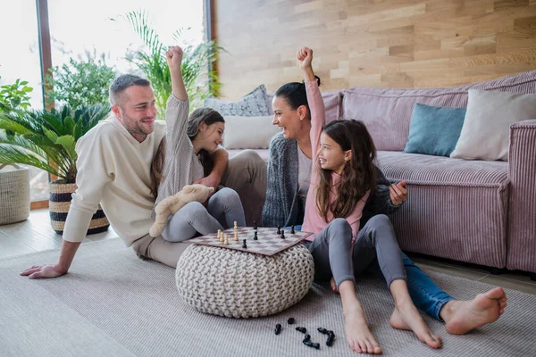 Duas irmãs felizes com mãe e pai sentados no chão e jogando xadrez juntos em casa — Fotografia de Stock