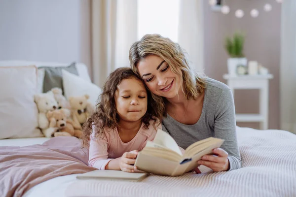 Счастливая мать с маленькой дочкой, лежащей на кровати и читающей книгу дома. — стоковое фото