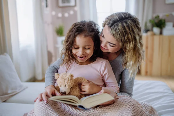 Счастливая мать со своей маленькой дочерью сидит на кровати и читает книгу дома. — стоковое фото