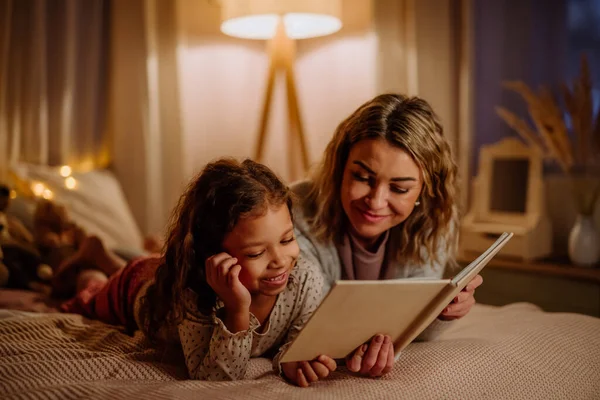 Madre feliz con su pequeña hija acostada en la cama y leyendo el libro por la noche en casa. — Foto de Stock