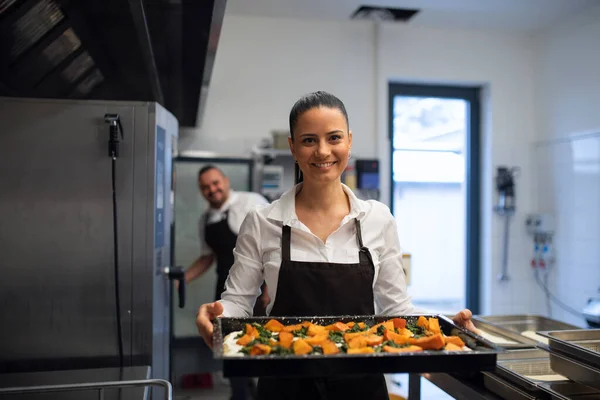 Happy female cook looking at camera and holding tray with baked pumpkin pieces in commercial kitchen — Stock Photo, Image