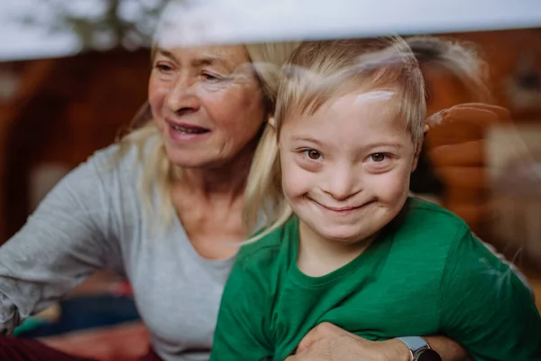 Мальчик с синдромом Дауна с бабушкой смотрит в камеру через окно дома. — стоковое фото