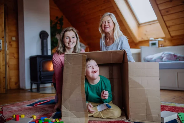 Junge mit Down-Syndrom mit Mutter und Großmutter spielt zu Hause mit Schachtel. — Stockfoto