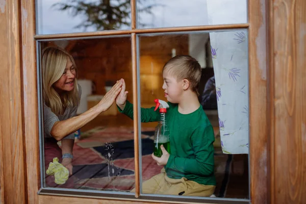 Niño con síndrome de Down con su abuela limpiando ventana en casa. — Foto de Stock