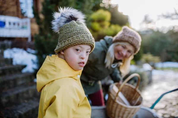 Down sendromlu çocuk kameraya bakıyor ve kışın büyükannesiyle bahçede çalışıyor.. — Stok fotoğraf