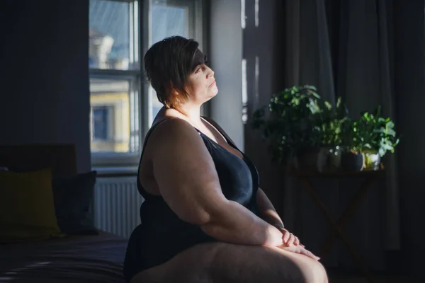 Deprimida mujer gorda sola sentada y mirando a través de la ventana en casa. — Foto de Stock