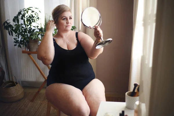 Kobieta z nadwagą siedząca i patrząca w lustro w domu, koncepcja samoopieki. — Zdjęcie stockowe