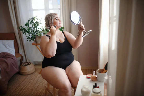 Mulher com excesso de peso sentada e olhando para o espelho e aplicando maquiagem em casa, conceito de autocuidado. — Fotografia de Stock