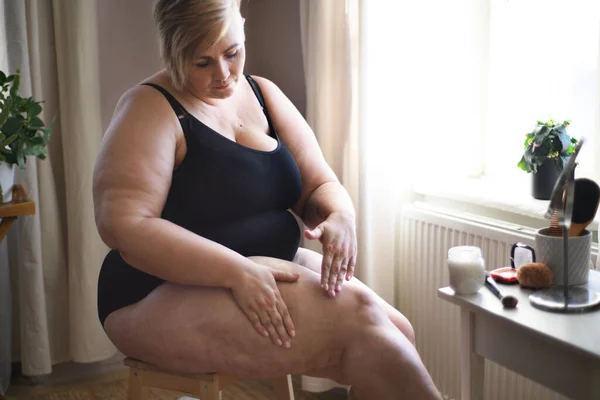 Mujer con sobrepeso sentada y aplicando crema en sus piernas en casa, concepto de autocuidado. — Foto de Stock