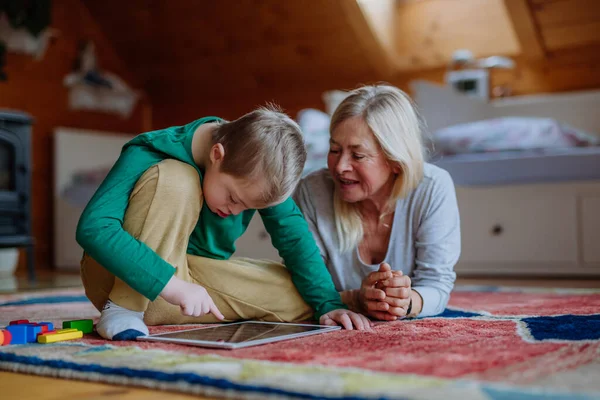 Junge mit Down-Syndrom mit seiner Großmutter, die auf dem Boden sitzt und zu Hause Tabletten nimmt. — Stockfoto