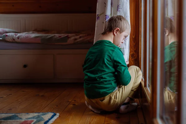 Niño pequeño con síndrome de Down sentado en el suelo y mirando a través de la ventana en casa. —  Fotos de Stock