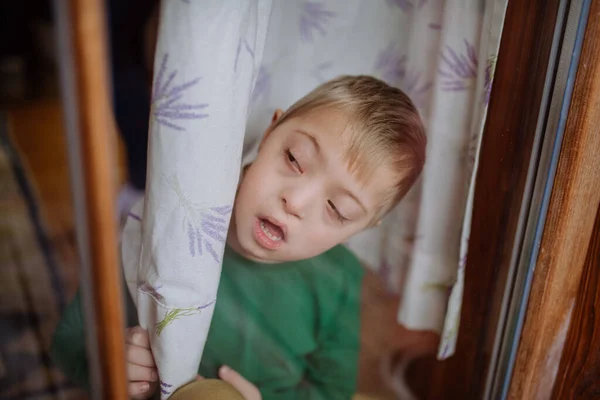 Kleiner Junge mit Down-Syndrom schaut zu Hause durchs Fenster. — Stockfoto