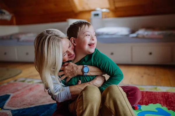 Happy boy met Down syndroom zitten op de vloer en knuffelen met zijn grootmoeder thuis — Stockfoto
