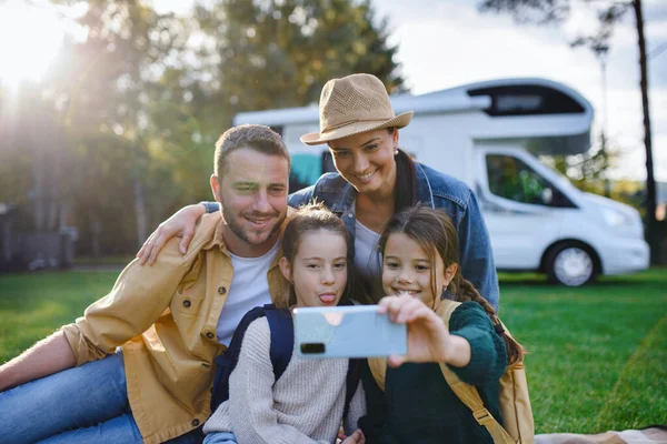 Šťastná mladá rodina se dvěma dětmi ltaking selfie s karavanou v pozadí venku. — Stock fotografie
