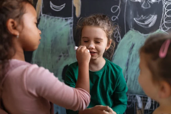 Маленькая девочка рисует мелом на щеках других девочек в помещении в игровой комнате. — стоковое фото