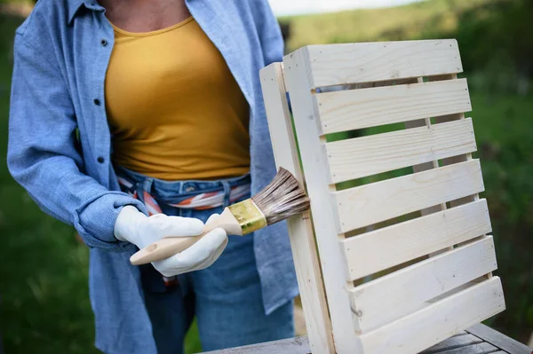 Close up of senior woman hand with paintbrush impregnating wooden crate outdoors in garden. — Stock Photo, Image