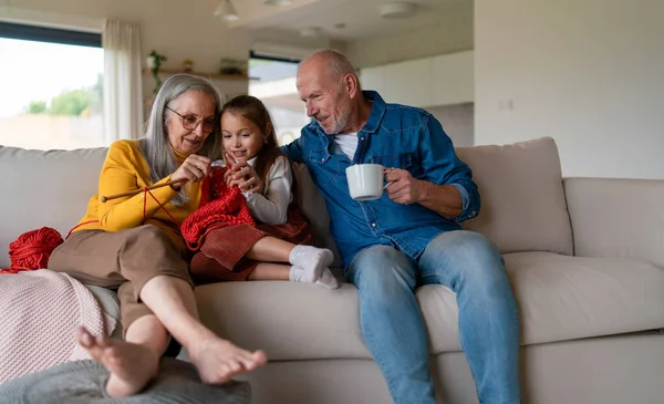 Маленькая девочка сидит на диване со своими бабушкой и дедушкой и учится вязать дома. — стоковое фото
