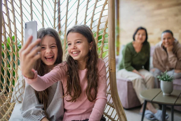 Gelukkig zusjes zitten in rieten rotan hang stoel binnen in serre en het nemen van selfie. — Stockfoto