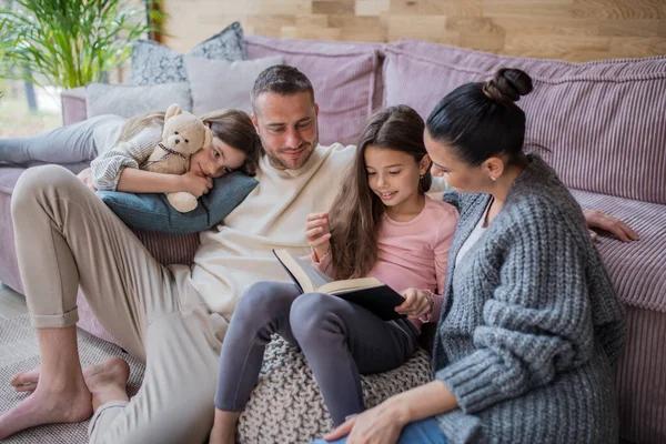 Due sorelle felici con madre e padre seduti sul pavimento che abbracciano e leggono il libro a casa — Foto Stock