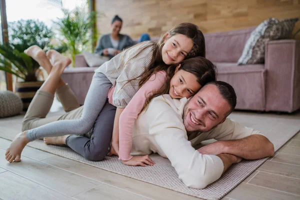 Två glada systrar har kul med far, ligger på rygg med mamma i bakgrunden hemma — Stockfoto